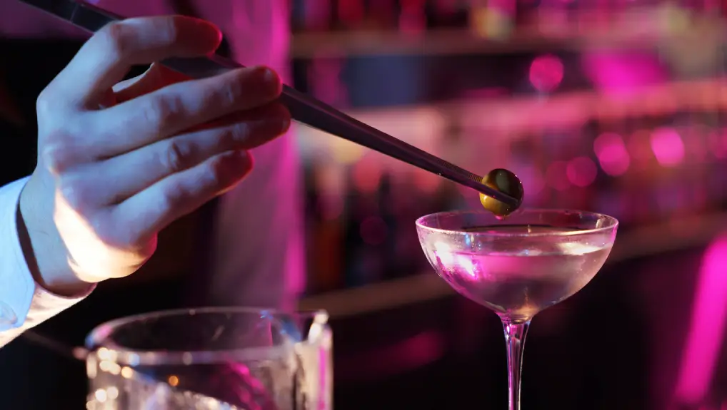 Bartender adding olive into Martini cocktail in bar- the martini glass story