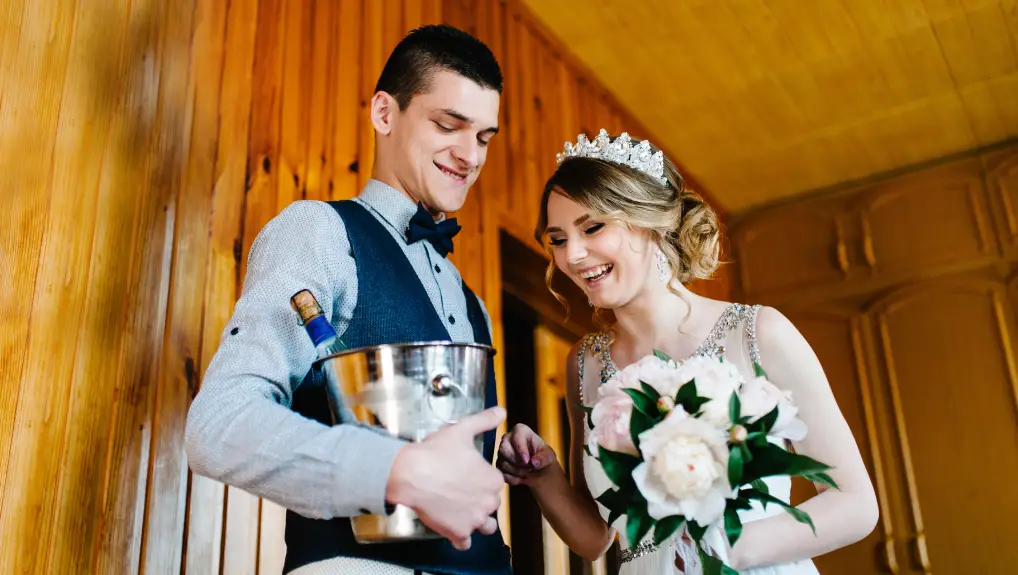 Image of a stylish happy groom with a butterfly keeps a bucket, pail of ice and champagne. Bride with a bouquet of flowers Peonies. On the background of a wooden wall and a picture. Wedding Morning. Close up
