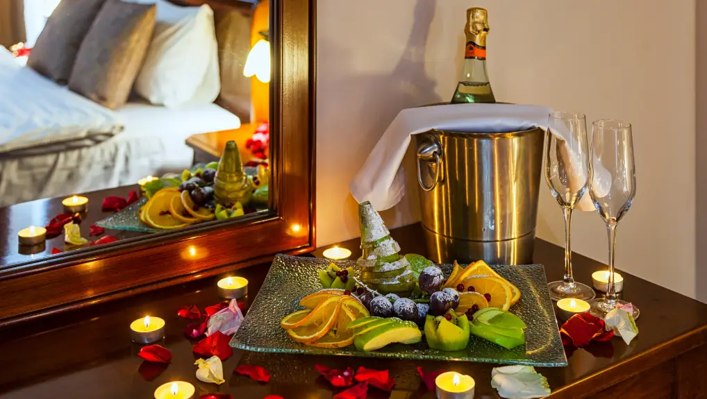 image of a table with a fruit plate, glasses of champagne, champagne with ice in a metal bucket and candles, in the background a bed decorated rose petals