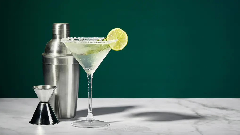 An image of Martini cocktail with ice and slice of lime, and shaker on a marble table. An alcoholic cocktail or non-alcoholic mocktail