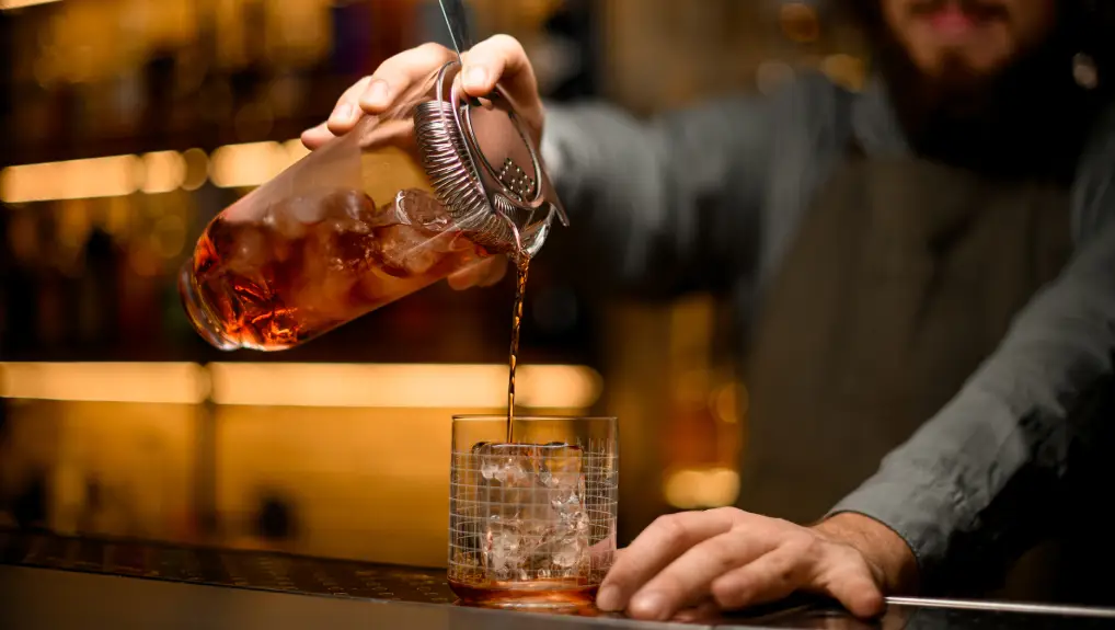 Image of a Hand of bartender holds transparent mixing cup with ice cubes and drink with strainer and poured drink into glass on bar