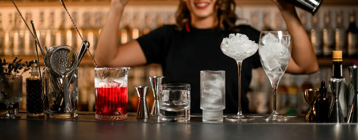 Image of a female bartender holding two cocktail shakers in front of various glasses and bartender tools on bar counter
