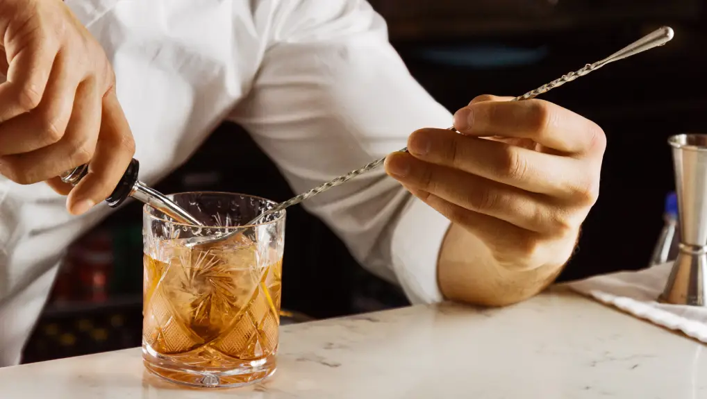 elegantly prepares a mixed drink by pouring all the ingredients into a bar spoon