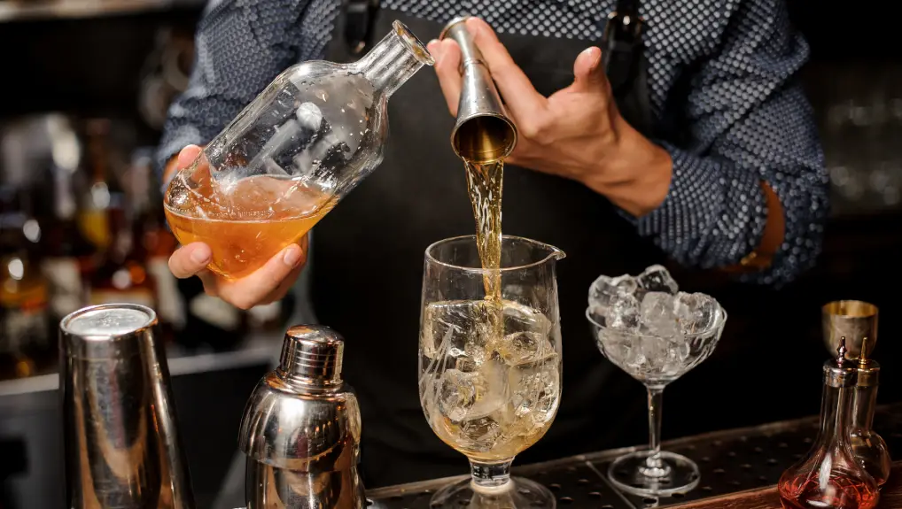 Image of a Barman pouring a portion of syrup from the bottle into the large cocktail glass with ice cubes on the bar counter