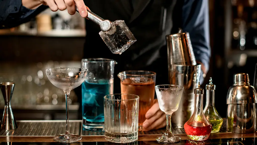 Image of Bartenders hand holds tongs with large square piece of ice over glasses