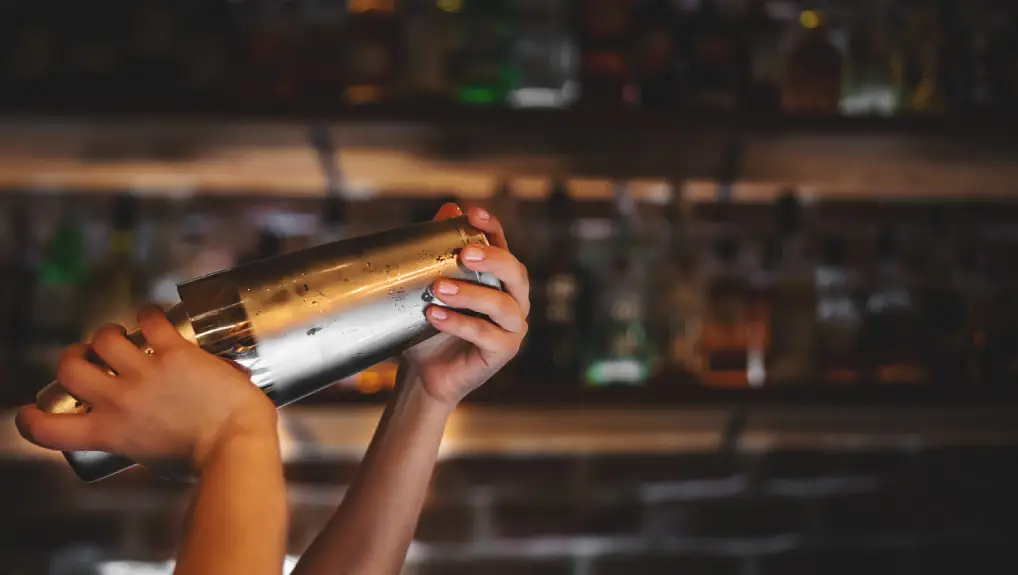 image of woman at bar holds steel shaker in her hands, shakes it to make cocktail.