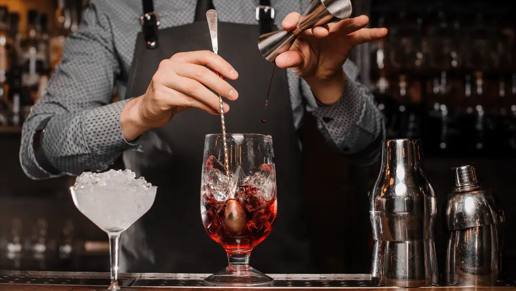 Image of arman in shirt and apron making an alcoholic drink with ice in a mixing glass
