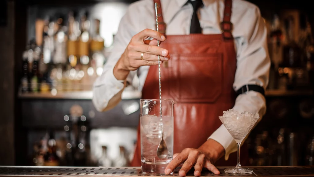 An image of man in apron stirring an ice cubes in the cocktail with bar spoon on the bar counter