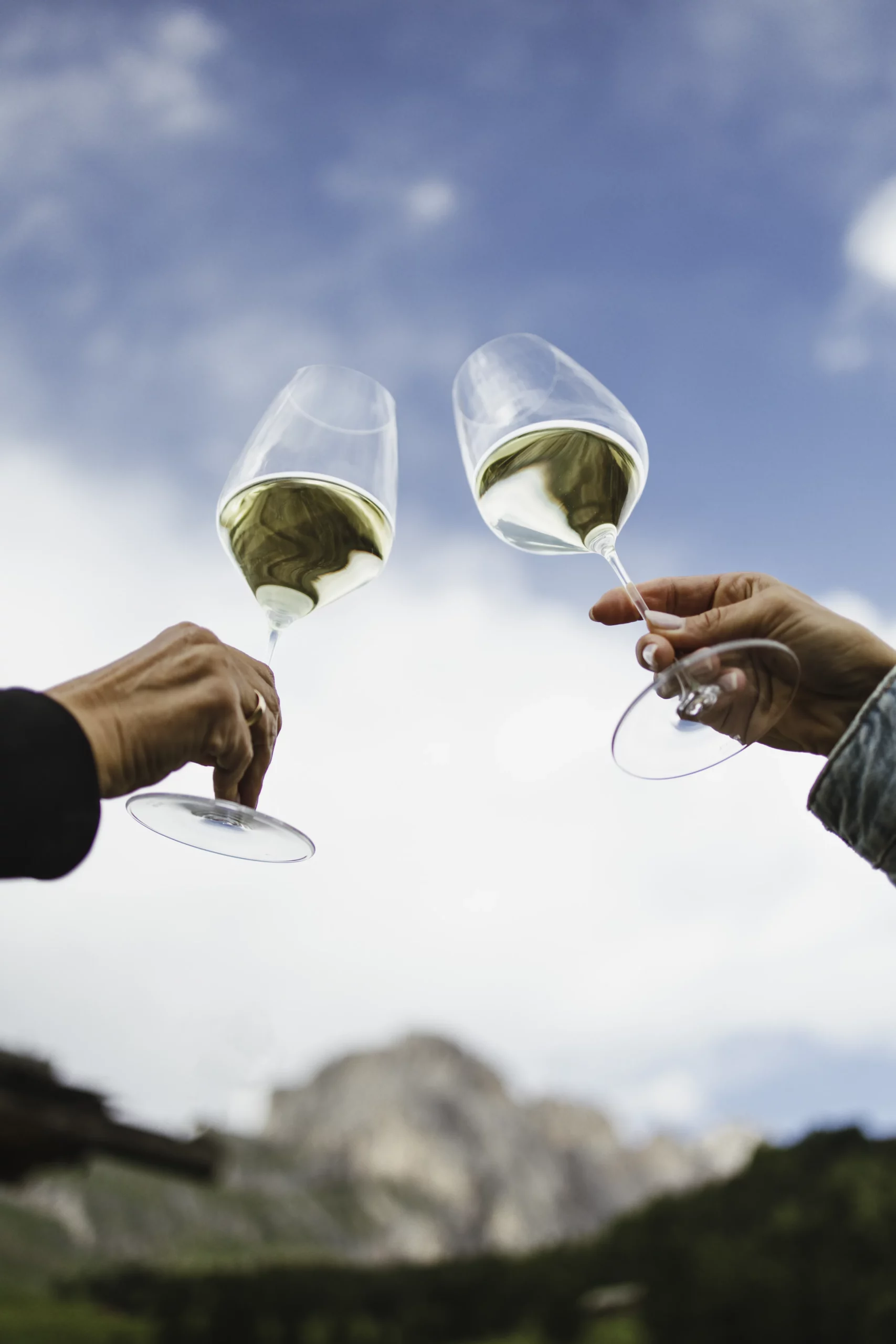 Two people's arms holding up wine glasses with a light blue sky with foggy clouds