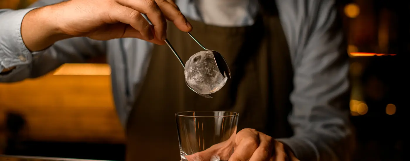 image of a bartender putting a sphere of ice into a whisky glass.