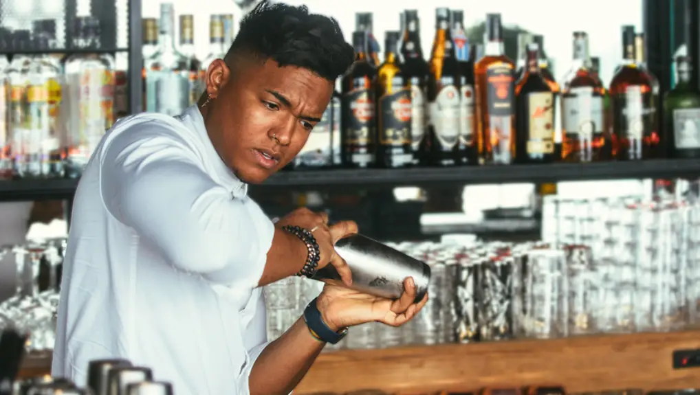 male expert barman is shaking a shaker for preparation of a cocktail at the bar counter- shows bar tools history