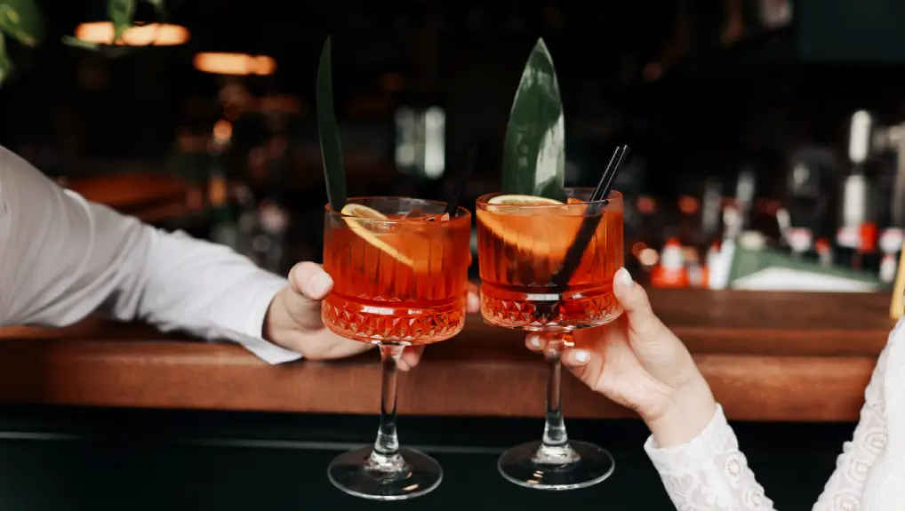 Image of hands of woman and man are clinking, cheers with glasses of Spritz cocktail. Couple celebrating wedding, anniversary with Spritz cocktails, with orange and greens Refreshing alcoholic drink