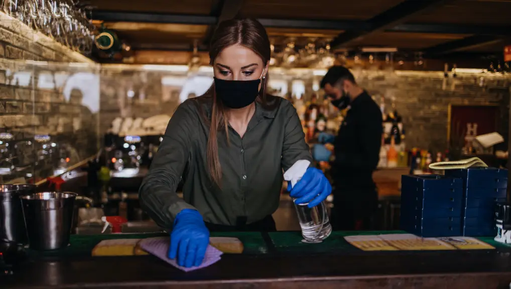 Portrait of a waitress standing behind a bar in a nice restaurant. She wears a protective mask and gloves as part of sanitising measures