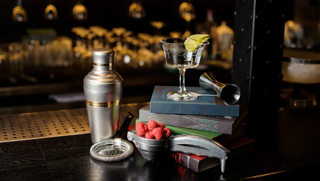 Image of Cocktail glass with lime slice and bar utensils arranged among books on the bar counter