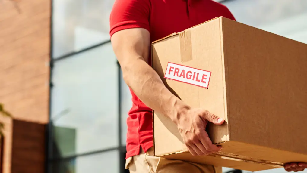 Image of delivery man carrying box and wearing red outdoors in sunlight in urban city setting 